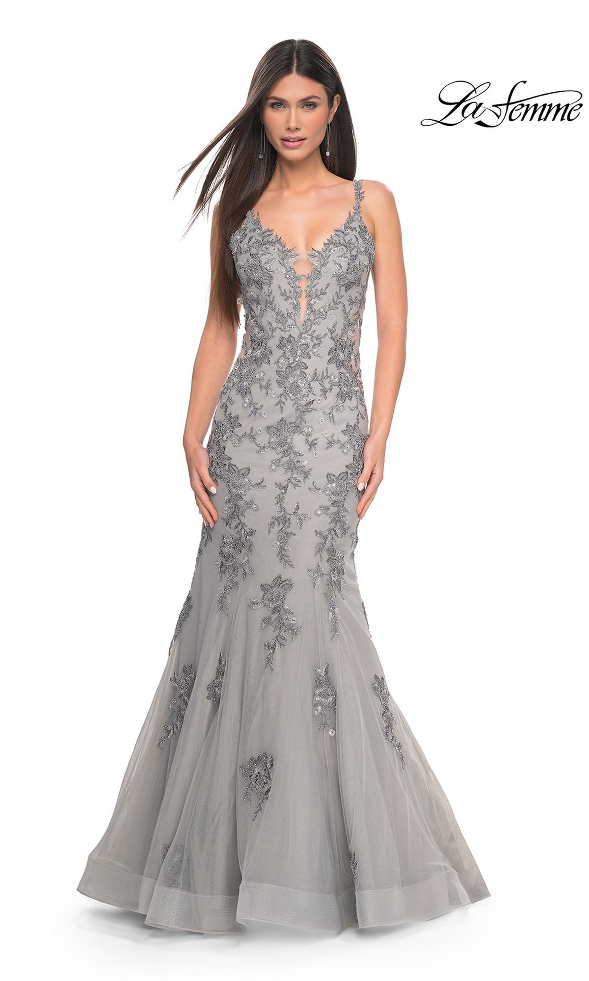 A woman in a floor-length, sleeveless silver La Femme Backless Long Mermaid Prom Dress 32295 with floral lace appliqués and a deep V neckline poses against a white background. The